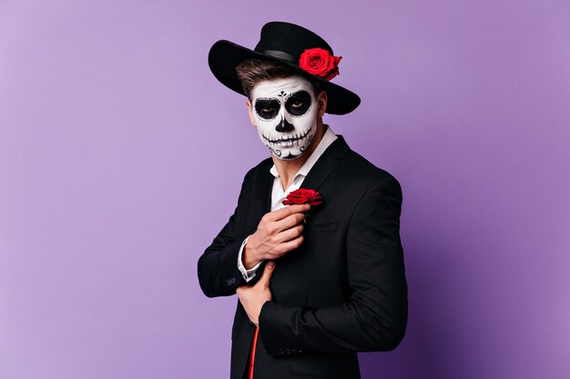 Zombie man in black hat posing on purple background. Indoor shot of male model in sombrero celebrating halloween.