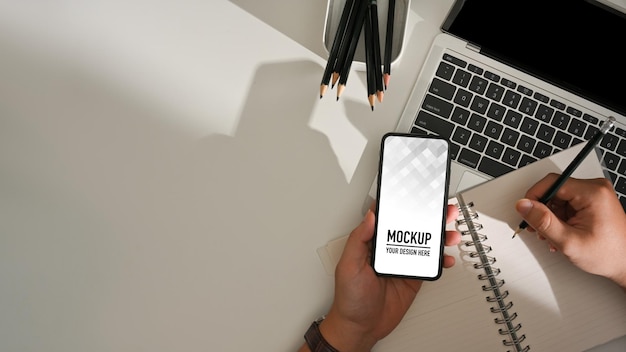 Top view of male hand writing on blank notebook and holding smartphone mockup