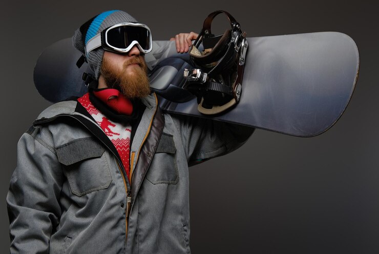 Gratis Foto a brutal man with a red beard wearing a full equipment holding a snowboard on his shoulder, isolated on a dark background.