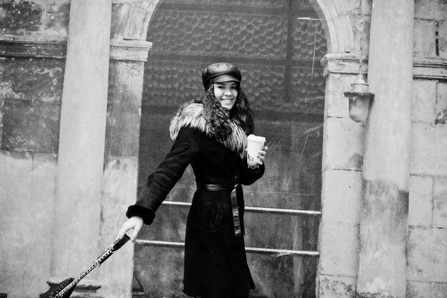curly mexican girl in leather cap and plastic cup of coffee at hand walking at streets of city