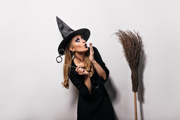 Gratis Foto funny caucasian girl posing in witch costume in carnival. long-haired woman in magic hat standing on white wall with broom.