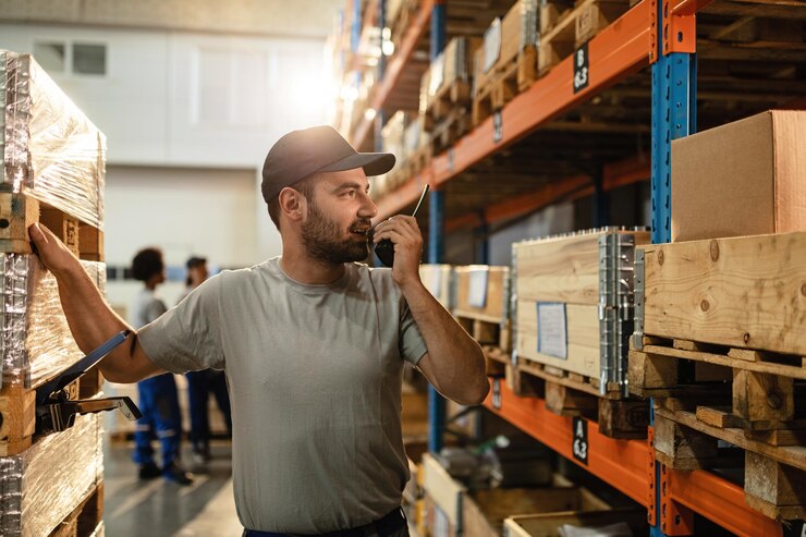 Gratis Foto male warehouse worker communicating with someone over walkietalkie while standing among shelfs in distribution warehouse