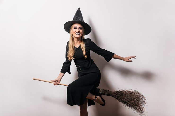 Gratis Foto søt heks i lang kjole som sitter på kosteskaft med et smil. bekymringsløs blond dame i karnevalskostyme som nyter halloween.