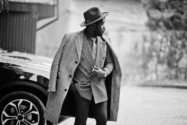 Stylish African American man model in gray coat jacket tie and red hat against black suv car Black and white photo