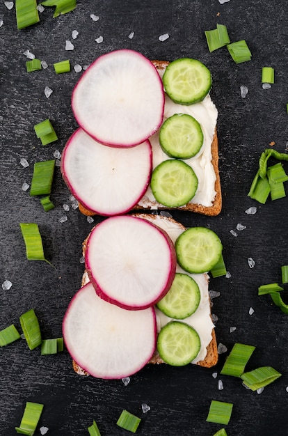 Top view of toasts with radish and cucumber on black surface vertical