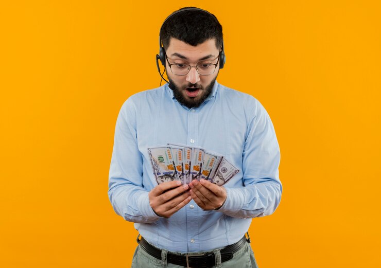 Gratis Foto young bearded man in blue shirt with headphones with microphone holding cash looking surprised and amazed standing over orange wall
