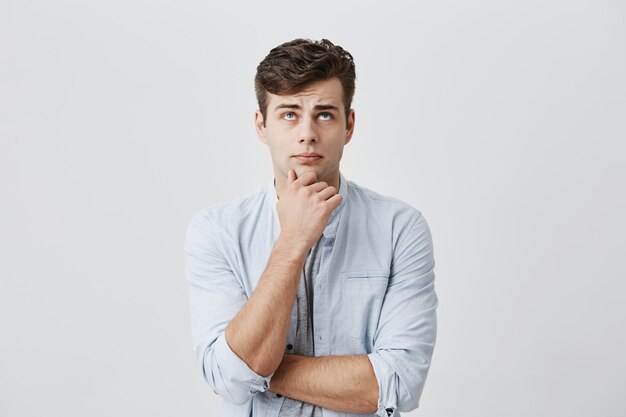 puzzled thoughtful male student dressed in light blue shirt, holding hand under his chin, frowning face, looking upwards, being dissatisfied with problems at university, thinking over his mistakes.
