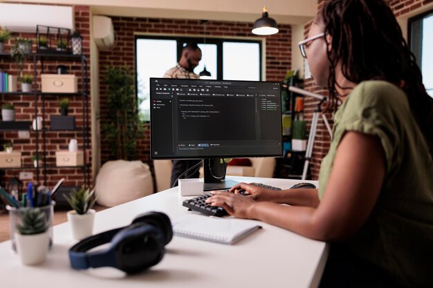 Woman coder developing software for freelance project working remote from home while boyfriend is relaxing. African american programmer writing source code looking for errors while debugging.