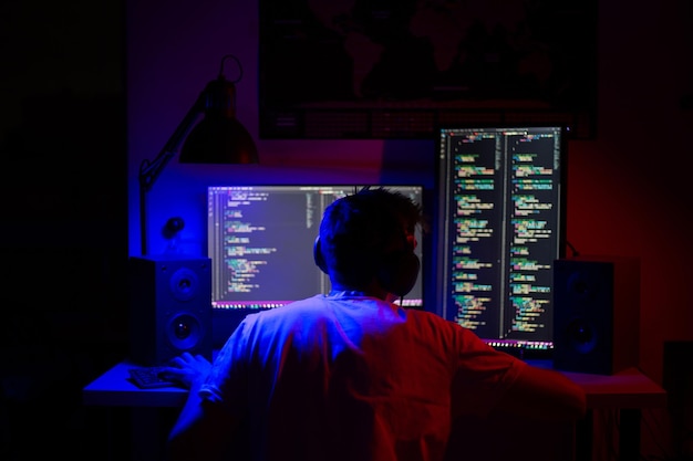 A man sits at a computer in a room at a table at night with blue lighting and programs