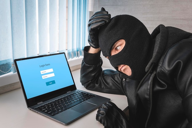 A thief in a balaclava mask is thinking about a password to log in The concept of identity theft Hacker With Log On Screen Computer Fraud Concept Background