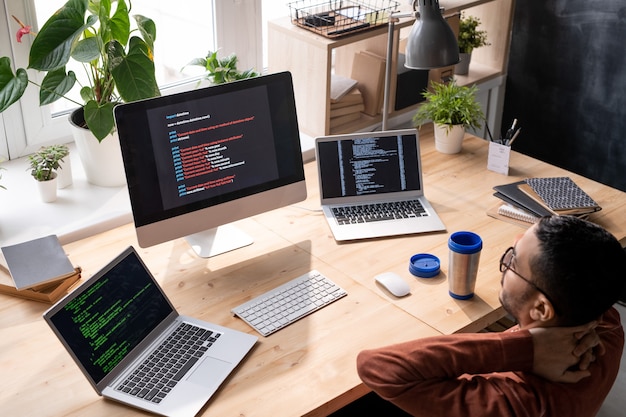 Above view of puzzled middle-eastern programmer in glasses sitting in front of computers and analyzing web script