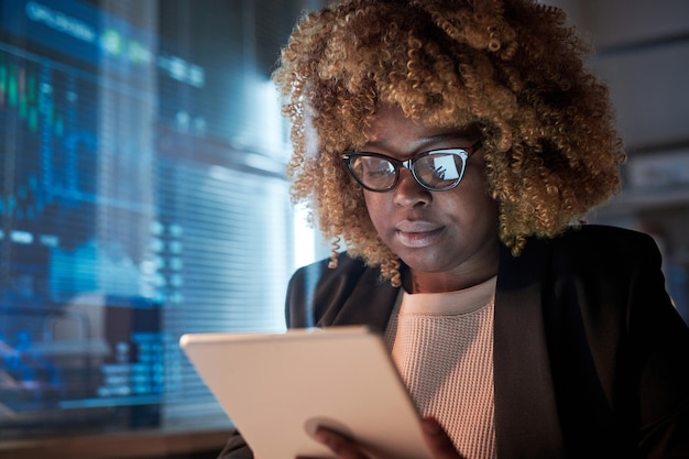 African female programmer with curly hair wearing eyeglasses developing software on digital tablet she working at office till late night