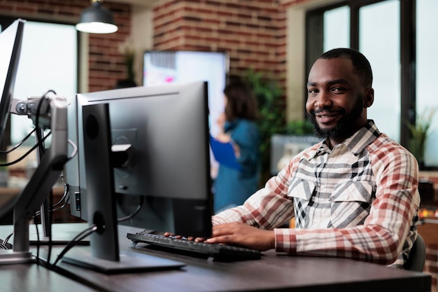 agency adult happy smiling worker at computer in startup studio space modern interior. african american creative company employee sitting at desk in office workspace while looking at camera.