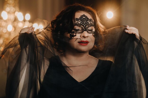 an elegant adult woman in a black cocktail dress and mask poses in the interior fashionable female portrait