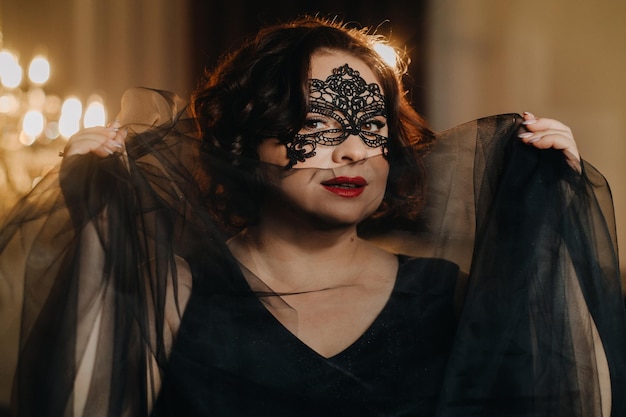 an elegant adult woman in a black cocktail dress and mask poses in the interior fashionable female