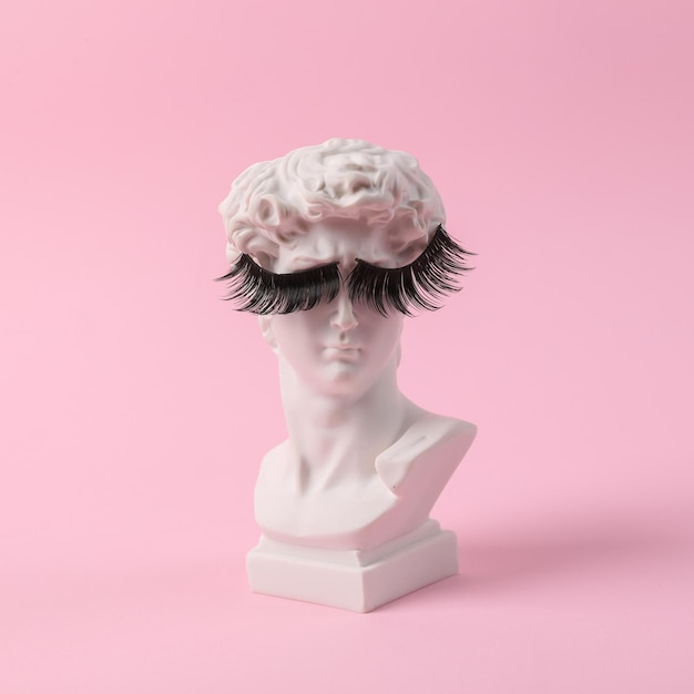 Antique bust of David with false eyelashes on a pink background Conceptual pop Minimal beauty and fashion still life Creative idea