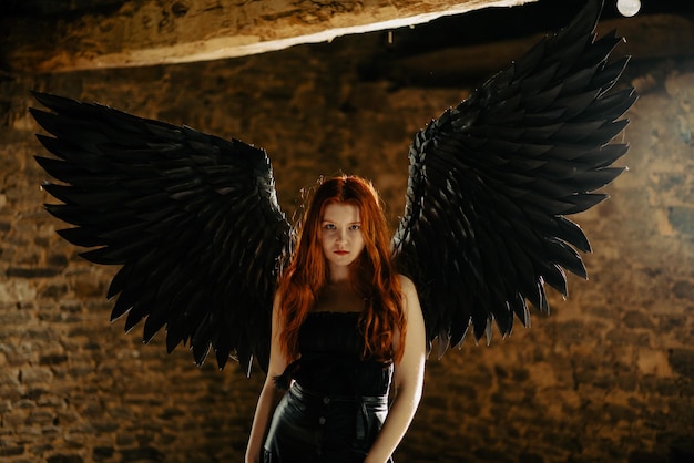beautiful young girl with long red hair and black wings in a dark room