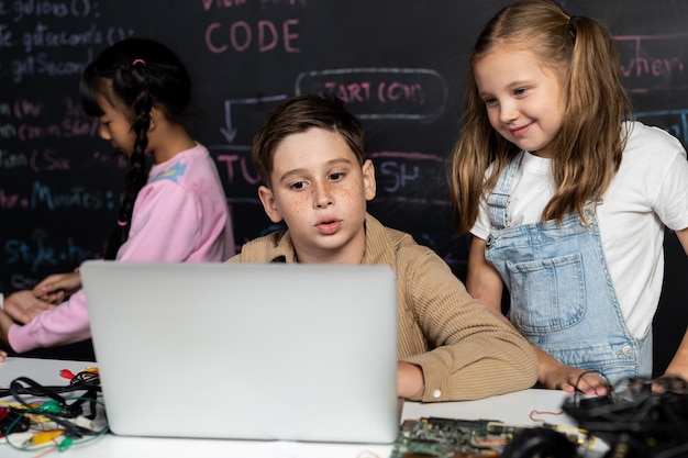 Brown shirt student and white bib student watch laptop and discuss Erudition