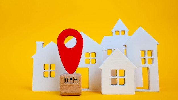 Cardboard boxes and a red position pin in city of white toy house on yellow background Locating packages and goods