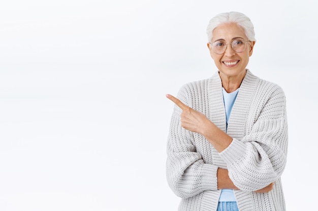 Cheerful pleasant senior woman, grandmother with grey combed hair, glasses, pointing upper left corner to promote advertisement