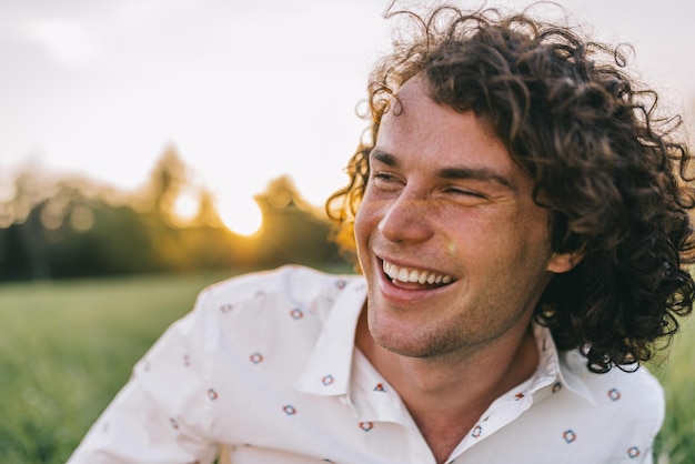 close up portrait of happy young male with curly hair smiling and relaxing on the green lawn in the park listening funny story copy space for advertising people and lifestyle concept