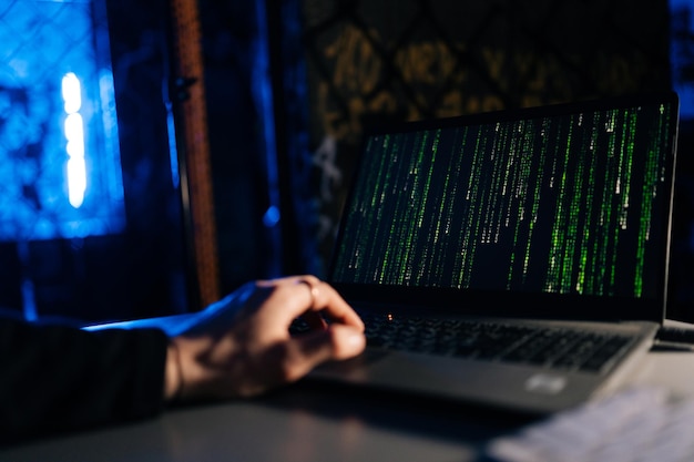Closeup of unrecognizable hacker engaged in hacking into security system on laptop green binary code on screen with sitting at desk in dark basement room