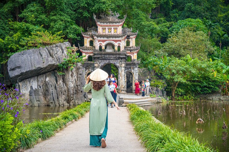 Photo cô gái mặc áo dài đứng từ phía sau, trước khung cảnh công viên ngoài trời với hồ nước và cổng cầu đá dẫn vào quần thể chùa bích động cổ kính, điểm đến du lịch ninh bình, việt nam