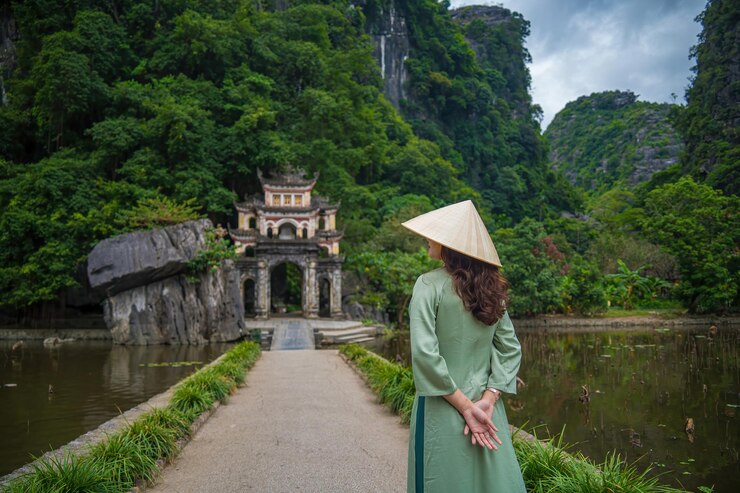 Photo cô gái mặc áo dài đứng từ phía sau, trước khung cảnh công viên ngoài trời với hồ nước và cổng cầu đá dẫn vào quần thể chùa bích động cổ kính, điểm đến du lịch ninh bình, việt nam