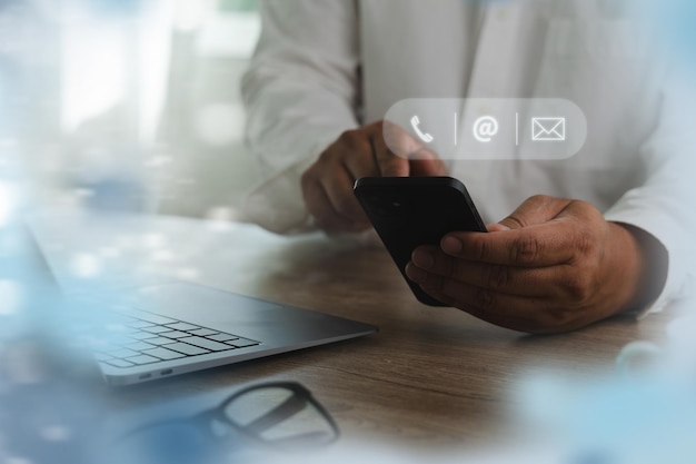 Contact us person using smartphone with digital icons for communication floating above sitting desk with laptop bright modern office environment