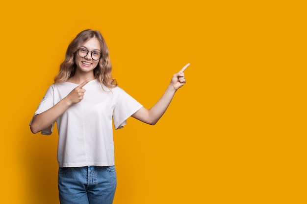 Curly haired woman with eyeglasses wearing white-shirt and jeans is advertising something by pointing to yellow free space near her