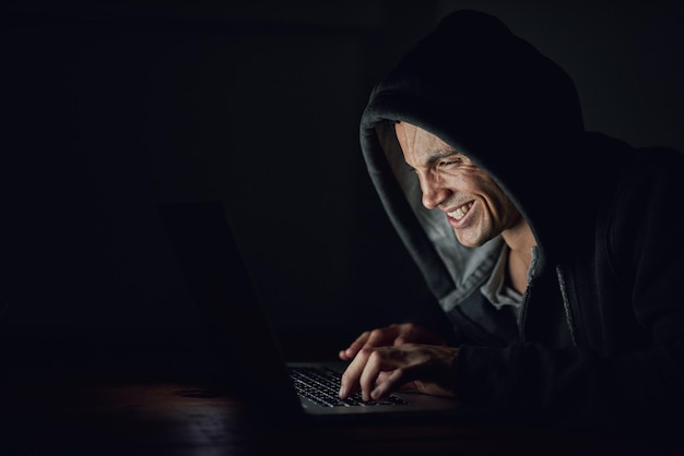 Dark basement and man at laptop for hacking online programming and cybersecurity at night Software developer or hacker in hoodie coding for password firewall or network access for cyber attack