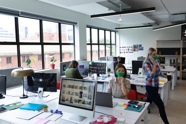 Diverse group of business people wearing face masks in creative office. group of people sitting at desks and using laptops. social distancing protection hygiene in workplace during covid 19 pandemic.