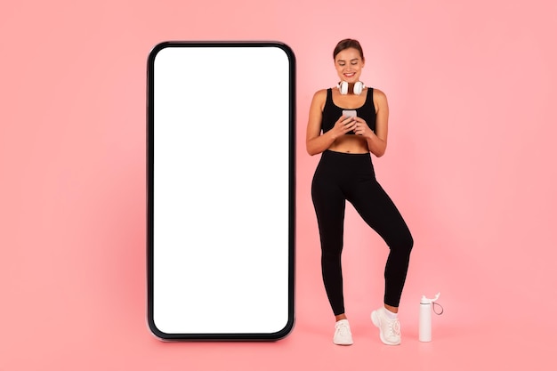 Happy Sporty Lady With Smartphone In Hands Standing Near Huge Blank Cellphone