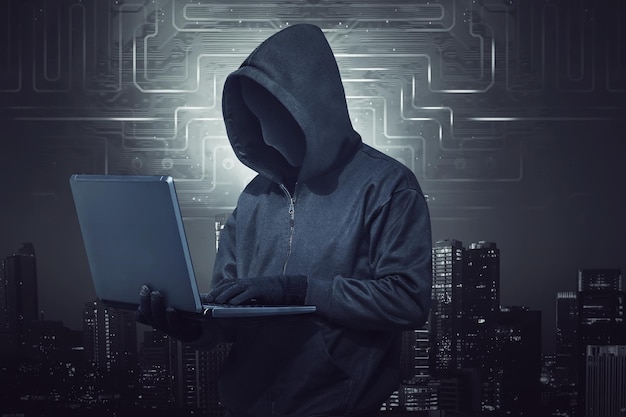 Hooded hacker holding laptop while typing against digital city background