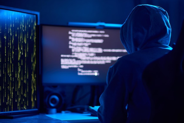 In a dimly lit room a cloaked figure types code on a computer hinting at a mysterious hacker
