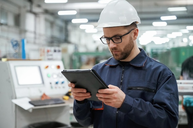 Looking and holding tablet Factory worker is indoors with hard hat