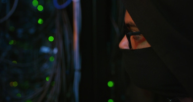 Night hacker and person with mask by cables for scam phishing or cybercrime for online attack Anonymous privacy and programmer with security breach server or database encryption in control room