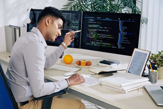 Pensive Indian developer checking structure chart when working on programming code at his desl