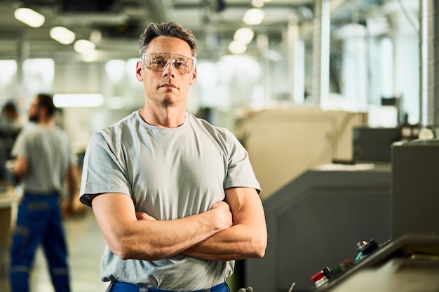 Portrait of confident engineer standing with arms crossed while working at CNC machine in industrial facility