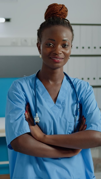 portrait of medical nurse looking at camera in cabinet standing in healthcare uniform with stethoscope. young person smiling while doctor and elderly patient talking in background