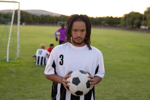 portrait of serious biracial male player in white uniform holding soccer ball standing at playground