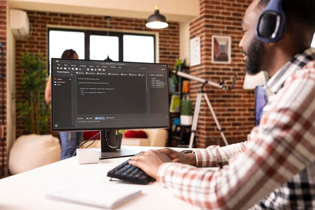 Remote male programmer sits at desk with headphones coding and debugging scripts on computer african