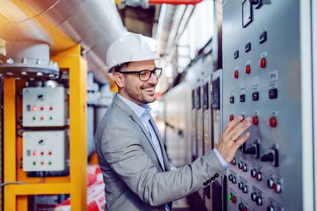 Smiling handsome caucasian supervisor in gray suit and with helmet on head turning switch on. Power plant interior.