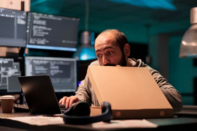 Software developer eating pizza from takeaway delivery, taking bite of food while he works after hours on source code development. Programmer having late dinner with takeout meal in carton box.