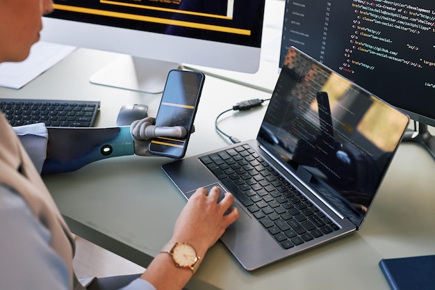 Software developer holding smartphone in bionic hand when programming in laptop