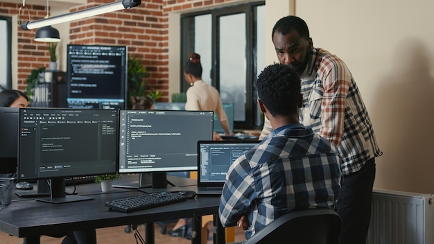 software developer writing code on laptop and computer keyboard looking at multiple screens with programming language is interrupted by coworker asking for advice. cloud programers doing teamwork.