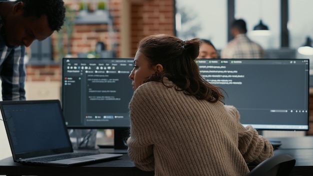 software programer putting laptop with source code on colleague coder desk asking for opinion about database. developer writing algorithm interrupted by coworker wanting help with fixing errors.
