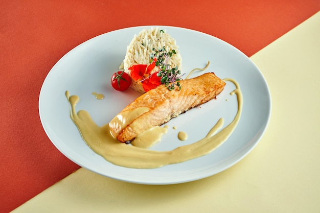 stir-fried salmon steak with yellow sauce and rice on a side dish in a white plate on colored surface.