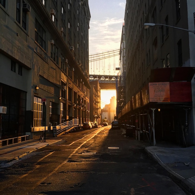 street amidst buildings leading towards manhattan bridge against sky during sunset