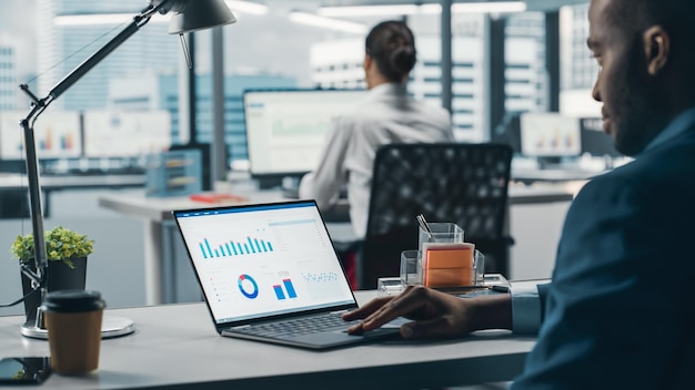 Successful Black Businessman Sitting at Desk Working on Laptop Computer in Big City Office Hard Working Top Manager Doing Research for eCommerce Project with Graps Charts and Tables Over Shoulder
